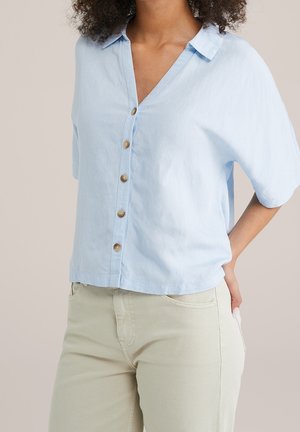 Mujer con camisa de manga corta azul claro abotonada y pantalones beige, de pie con una mano en la cadera sobre un fondo liso.