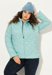 Giacca trapuntata azzurro chiaro con colletto alto e zip, caratterizzata da un motivo geometrico. Abbinata a pantaloni scuri a fantasia e un berretto blu.