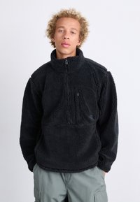 Pull en polaire noir avec un col montant, poche zippée sur la poitrine, surface texturée et poignets élastiques. Présente le logo de la marque à l'avant.