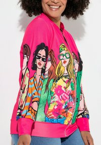 Lyserød zip-up jakke med et farverigt grafisk print af forskellige karakterer. Designet inkluderer dristige mønstre og kontrasterende kanter.