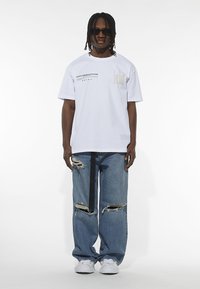 White short-sleeve T-shirt featuring printed text; paired with loose-fitting, distressed blue jeans. Worn with sunglasses and white shoes.