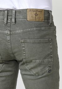 Gros plan sur le dos d’un jean en denim gris avec un patch de marque en cuir carré, une poche arrière visible et des passants de ceinture.