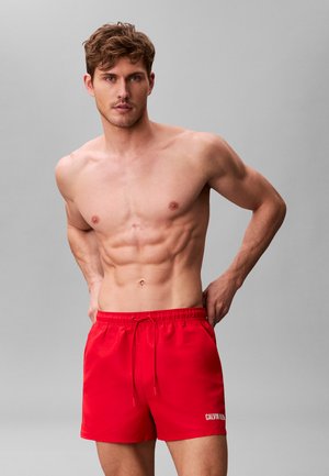 Skjorteløs mand med muskuløs krop, der justerer røde Calvin Klein badeshorts mod en ensartet grå baggrund.