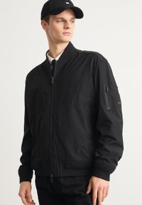 Černá bombardérka s předním zipem, žebrovanými manžetami a náprsní kapsou na rukávu. Hladká textura a strukturovaný design.