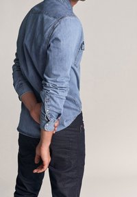 Camicia in denim azzurro chiaro con colletto button-down e maniche lunghe, caratterizzata da una tasca sul petto e polsini risvoltati, abbinata a jeans scuri.