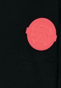 Czarna dzianina z wyraźną okrągłą gumową łatą w jasnoróżowym kolorze, z wytłoczonymi literami i ozdobną ramką.