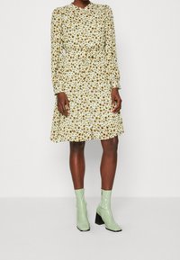 Abito floreale con maniche lunghe, aderente in vita, di un delicato crema con fiori marroni; abbinato a stivaletti verdi alla caviglia e tacchi a blocco.