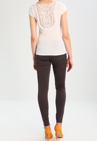 Biała bluzka z krótkim rękawem z koronkowym, kwiatowym tyłem, połączona z ciemnobrązowymi dopasowanymi spodniami oraz pomarańczowymi szpilkami.