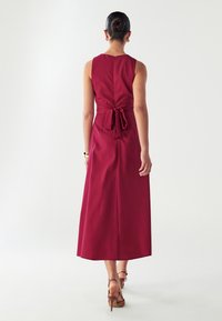 Burgundijska haljina bez rukava s zavezanom strukom, detaljem za vezanje na leđima i dugačkom, lepršavom suknjom. Glatka tkanina i čiste linije.