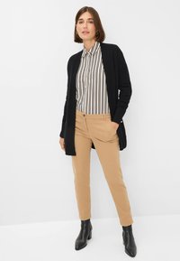 Cardigan en tricot noir sur une chemise à rayures blanches et noires, associé à un pantalon en toile beige et des bottines noires à talons. Tissu lisse, design ajusté.