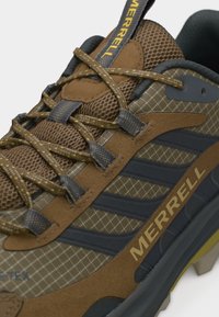 Καφέ και μαύρο πεζοπορικό παπούτσι Merrell με υφασμάτινο σχέδιο, κορδόνια με κίτρινες λεπτομέρειες και εμφανές λογότυπο στο πλάι και στη γλώσσα του παπουτσιού.