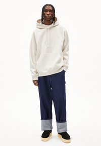 Felpa con cappuccio crema con tasca a marsupio, abbinata a jeans blu scuro di taglio ampio arrotolati alle caviglie e scarpe nere con suole spesse.