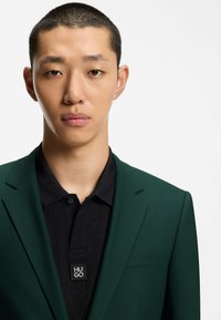 Junger Mann mit kurzen Haaren, der ein dunkelgrünes Jackett über einem schwarzen Poloshirt mit einem "HUGO"-Logo-Patch trägt.