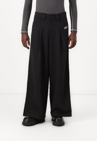 Muž v černých širokých kalhotách, černých botách s masivní podrážkou a upnutém šedém dlouhém rukávu s bílým prošíváním na rukávech.