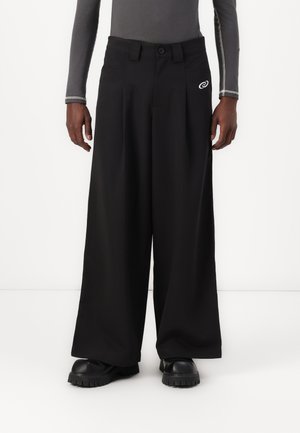 Man som bär svarta vida byxor, svarta klumpedans-skor och en figurnära grå långärmad tröja med vit sömnad på ärmarna.