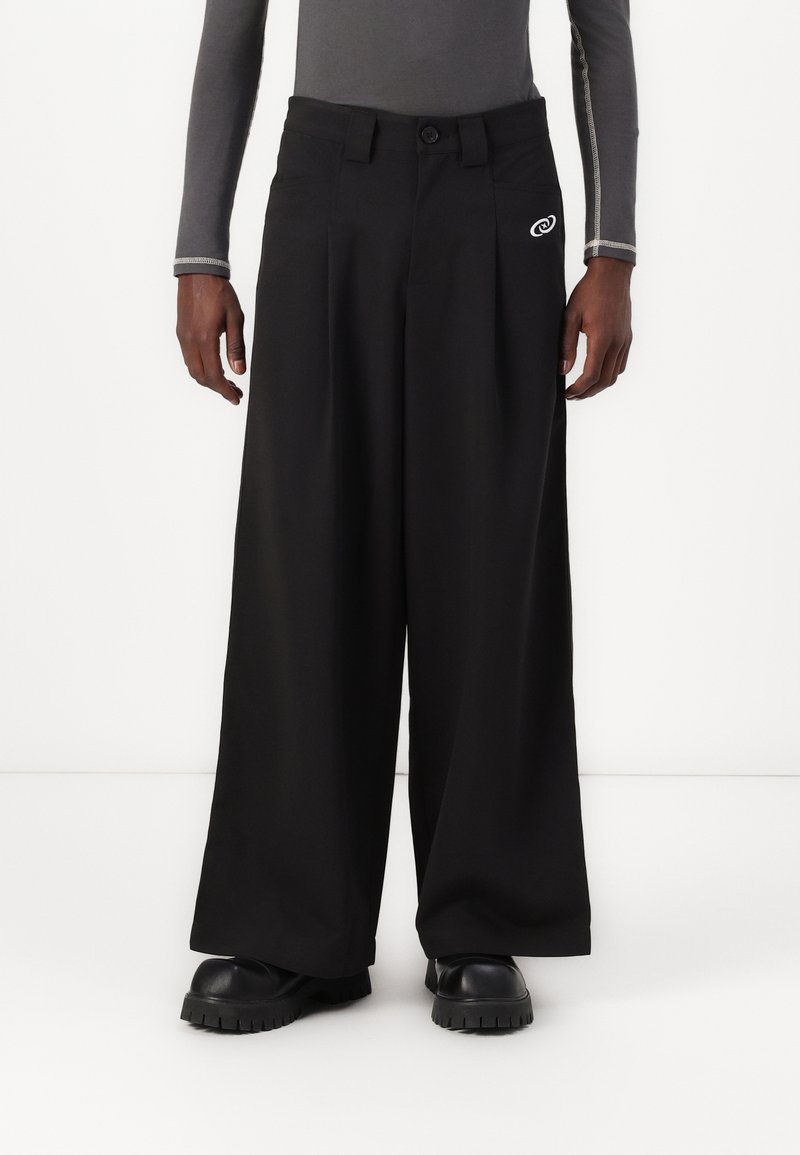 Muž v černých širokých kalhotách, černých botách s masivní podrážkou a upnutém šedém dlouhém rukávu s bílým prošíváním na rukávech.