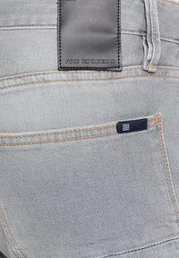 Lichtgrijze denim jeans met een achterzak met een marineblauw label. Inclusief leren taillebandpatch in zwart met geperst tekst "NO EXCESS."