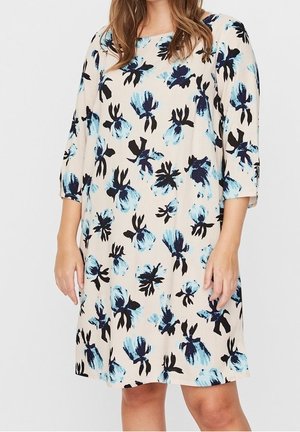 Vrouw draagt een beige jurk met blauwe en zwarte abstracte bloemenpatronen en driekwart mouwen.