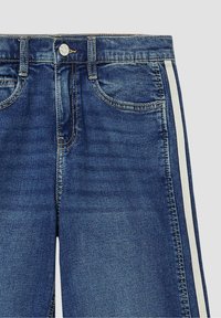 Jean denim bleu avec poches avant, bouton visible, fermeture éclair et bandes blanches sur les coutures extérieures, sur un fond uni.