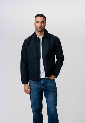 Mężczyzna ubrany w granatową bluzę na zamek, pod którą ma białą koszulę i niebieskie dżinsy, stoi z jedną ręką w kieszeni na jednolitym jasnym tle.