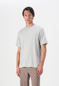 Tricou din bumbac gri, cu mâneci scurte și guler rotund, având o croială lejeră. Modelul poartă pantaloni maro.