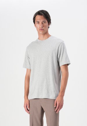 Grå bomulls-t-shirt med korta ärmar och rund halsringning, med en avslappnad passform. Modellen bär bruna byxor.