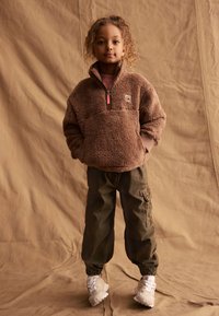 Brun fleece-tröja med dragkedja, khaki cargo-byxor med sidfickor och beige sneakers. Bakgrunden är strukturerad och neutralt tonad.