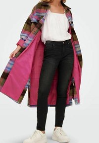 Woman in black skinny jeans, white sneakers, white sleeveless top, and open multicolor plaid coat with bright pink lining.
