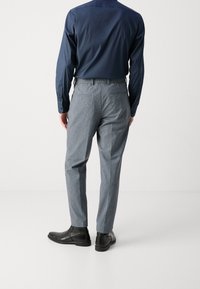 Muž v modré košili s dlouhým rukávem, šedých vzorovaných kalhotách a černých kožených společenských botách, stojící na bílém pozadí a otočený zády.