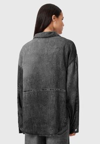 Donna con lunghi capelli scuri indossa una giacca di jeans nera over-size con effetto acidato, vista di spalle contro uno sfondo grigio chiaro semplice.