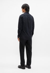 Cămașă neagră cu mânecă lungă, cu guler ascuțit, manșete cu nasturi și un tiv drept. Asortată cu pantaloni negri și pantofi negri, văzută din spate.