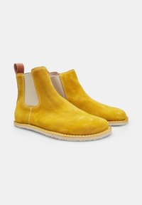 Botas Chelsea de ante amarillas con paneles laterales elásticos y una lengüeta de cuero beige, que presentan una superficie texturizada y una suela blanca contrastante.