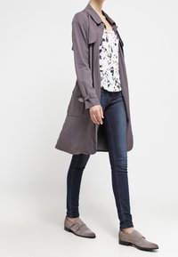 Jasnoszary trencz z dużym kołnierzem i przednimi kieszeniami, noszony na wzorzystej bluzce, w zestawie z ciemnymi dżinsami i szarymi butami.