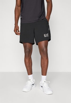 Pantaloni sportivi neri con un logo, due tasche laterali e una superficie liscia. Indossati con una maglietta nera e scarpe da ginnastica bianche.
