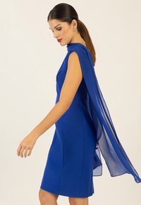 Blaues Kleid mit figurschmeichelndem Design, das mit einem fließenden Chiffon-Cape über den Schultern versehen ist, glatter Textur und einer Länge bis oberhalb des Knies.