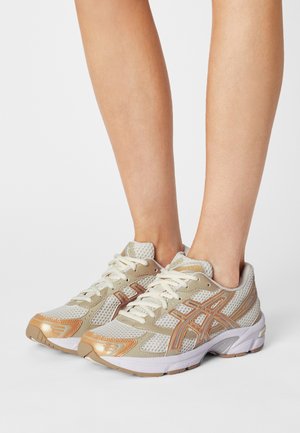 Béžové běžecké boty s síťovinou a syntetickým svrškem, smetanovými tkaničkami a metalickými akcenty. Bílá podrážka s mírným zakřivením a texturovaným vzorem.