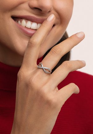 Anillo de plata con un diseño entrelazado que presenta una fila de pequeñas piedras. Superficie brillante y textura suave, llevado en una mano cerca del rostro.