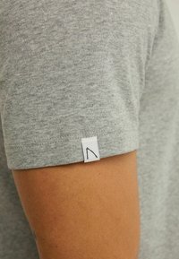 Tricou gri deschis, fabricat din bumbac moale. Prezintă o etichetă mică albă cu un logo conturat negru pe mânecă stângă. Design cu mâneci scurte.
