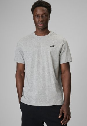 Mężczyzna ubrany w jasnoszary krótki t-shirt z małym logo 4F na piersi i czarne spodnie, stojący na jednolitym szarym tle.