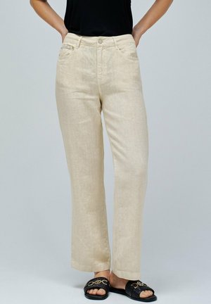 Pantalones de lino beige con corte recto, cierre de botón y bolsillos. Se llevan con sandalias negras que cuentan con una tira decorativa.