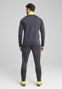 Idræts tracksuit i mørkegrå stof med høj krave, der har en klar neon gul detalje. Slim fit med lange ærmer og tætsiddende bukser.