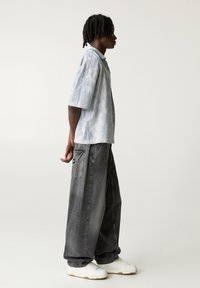 Chemise à manches courtes, de couleur bleu clair avec un motif tie-dye, assortie à un jean large gris foncé avec de grandes poches et une étiquette noire. Baskets blanches.