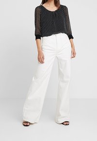 Schwarze, durchsichtige Bluse mit weißen Punkten, langen Ärmeln und schwarzen Bündchen, kombiniert mit hoch taillierten, weit geschnittenen weißen Hosen und schwarzen Sandalen.