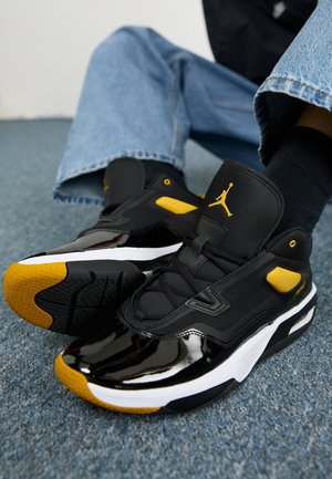 Sorte og gule atletikskøjter med en blank lakfinish, polstret krave, mesh-detaljer, gummisål og kontrasterende hvid mellemsål.