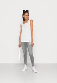Top sem mangas branco com padrão floral multicolorido, combinado com jeans justos cinzentos e ténis brancos, segurando uma pequena bolsa branca.