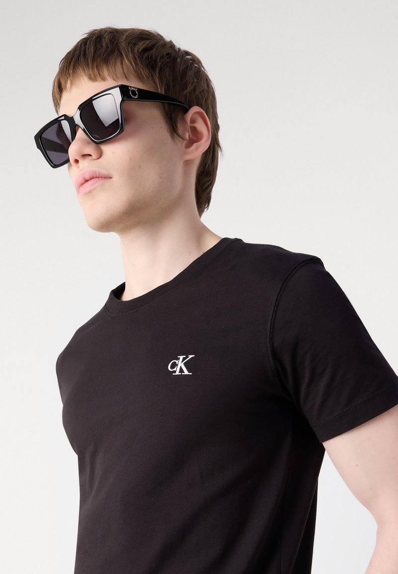 Joven con camiseta negra de Calvin Klein y grandes gafas de sol negras cuadradas, mirando hacia la izquierda con expresión neutra sobre fondo liso.