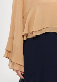 Personne portant un haut beige à manches à volants superposé à une jupe bleu marine foncé, une main visible sur le côté, sur un fond uni.