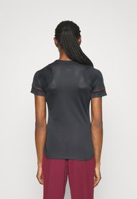 Person med långt flätat hår som är bundet, iklädd en tight mörkgrå kortärmad tröja och vinröda byxor med röda sidostråk, vänd bort.