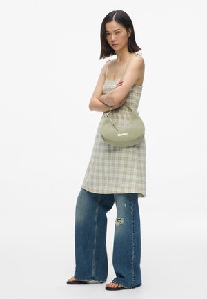 Woman in sleeveless plaid dress over ripped wide-leg jeans, black sandals, carrying light green shoulder bag, standing with arms crossed.