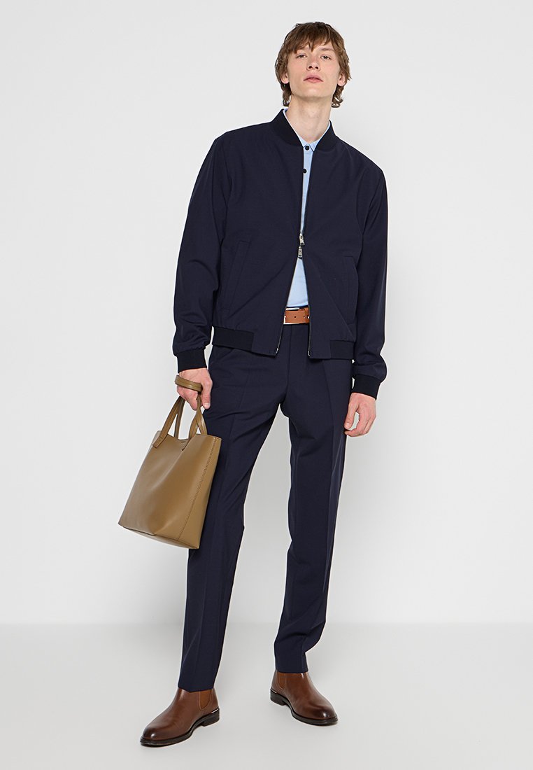 Marineblaues Bomberjacke über einem hellblauen Hemd, kombiniert mit dunklen Hosen, einem braunen Gürtel und braunen Lederschuhen. Hält eine beige Tote-Tasche.